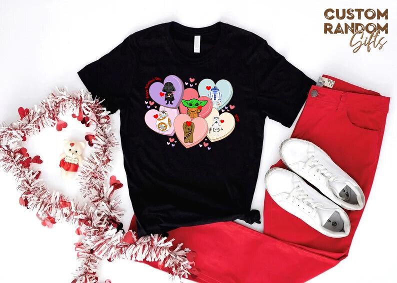 May include: Black t-shirt featuring Star Wars characters on heart-shaped candies. The design includes Darth Vader, Baby Yoda, R2-D2, BB-8, Chewbacca, and a Stormtrooper. Paired with red pants and white sneakers, with a Valentine's Day wreath.