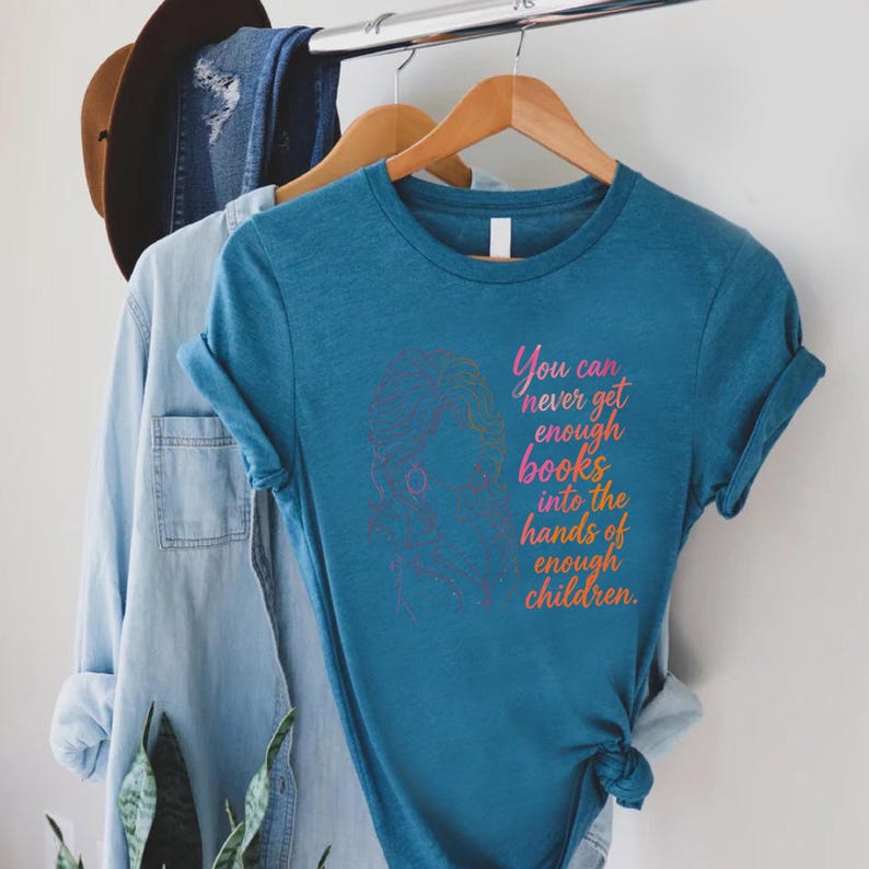 May include: A teal short-sleeved t-shirt featuring a line art illustration of a person with voluminous hair and the text, You can never get enough books into the hands of enough children, printed in a gradient of pink, orange, and yellow colors.