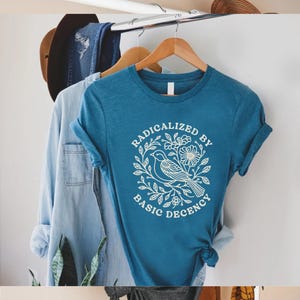 May include: Teal-colored t-shirt with rolled-up sleeves, featuring a white graphic of a bird and floral design, encircled by the words "RADICALIZED BY BASIC DECENCY." Other clothing items hang in the background.