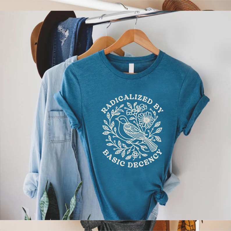 May include: Teal-colored t-shirt with rolled-up sleeves, featuring a white graphic of a bird and floral design, encircled by the words "RADICALIZED BY BASIC DECENCY." Other clothing items hang in the background.