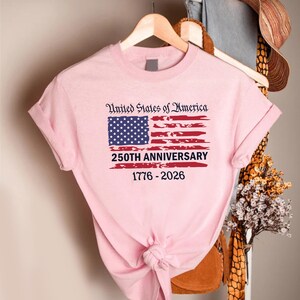 May include: A light pink t-shirt featuring the American flag with the text "United States of America" above the flag. Below the flag is the text "250TH ANNIVERSARY 1776-2026". The shirt is hanging on a wooden hanger.
