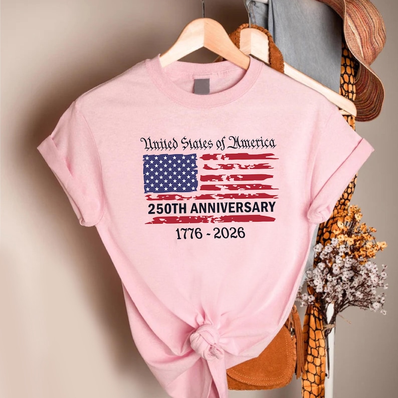 May include: A light pink t-shirt featuring the American flag with the text "United States of America" above the flag. Below the flag is the text "250TH ANNIVERSARY 1776-2026". The shirt is hanging on a wooden hanger.