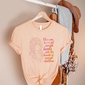 May include: A peach-colored short-sleeve t-shirt featuring a line-art illustration of a person with voluminous hair and the text, You can never get enough books into the hands of enough children, printed in a gradient of pink and orange ink.