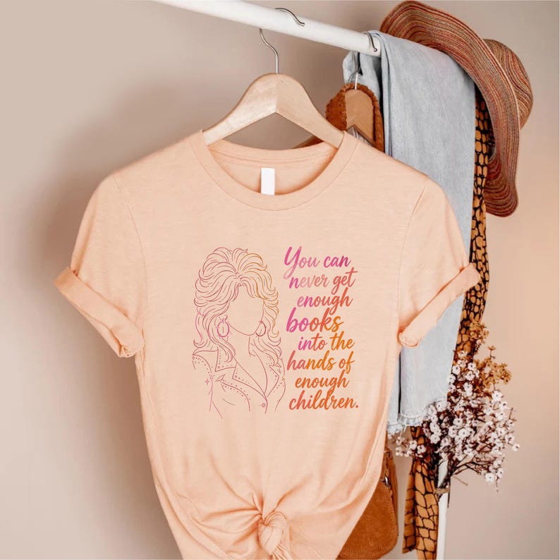 May include: A peach-colored short-sleeve t-shirt featuring a line-art illustration of a person with voluminous hair and the text, You can never get enough books into the hands of enough children, printed in a gradient of pink and orange ink.