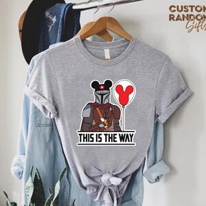 Puede incluir: Camiseta gris con una ilustraci&oacute;n de dibujos animados de un personaje mandaloriano con una cabeza de Mickey Mouse y un globo rojo. El texto "THIS IS THE WAY" est&aacute; impreso debajo del personaje en un cuadro blanco.