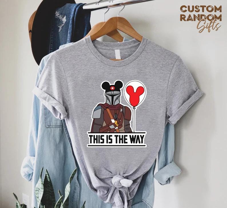 Puede incluir: Camiseta gris con una ilustraci&oacute;n de dibujos animados de un personaje mandaloriano con una cabeza de Mickey Mouse y un globo rojo. El texto "THIS IS THE WAY" est&aacute; impreso debajo del personaje en un cuadro blanco.
