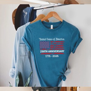 May include: A teal t-shirt with the text "United States of America" above a stylized American flag graphic. Below the flag, the text reads "250TH ANNIVERSARY 1776-2026". The shirt is hanging on a wooden hanger.