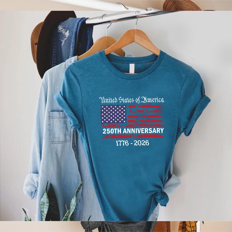 May include: A teal t-shirt with the text "United States of America" above a stylized American flag graphic. Below the flag, the text reads "250TH ANNIVERSARY 1776-2026". The shirt is hanging on a wooden hanger.