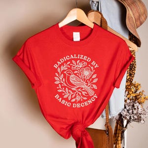 May include: A red t-shirt with a white graphic featuring a bird, flowers, and the text "RADICALIZED BY BASIC DECENCY." The shirt is hanging on a wooden hanger. The sleeves are cuffed.
