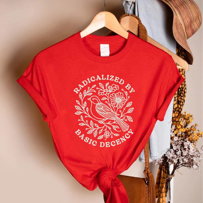 May include: A red t-shirt with a white graphic featuring a bird, flowers, and the text "RADICALIZED BY BASIC DECENCY." The shirt is hanging on a wooden hanger. The sleeves are cuffed.