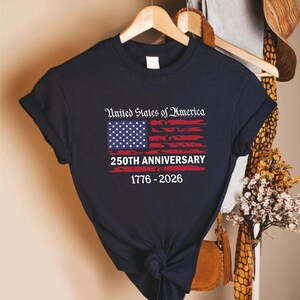 May include: Navy blue t-shirt featuring the American flag design with the text "United States of America 250th Anniversary 1776-2026". The flag design includes stars and stripes in red, white, and blue.