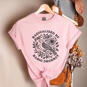 May include: A light pink t-shirt with rolled-up sleeves. The shirt features a black graphic of a bird surrounded by leaves and flowers, with the words "RADICALIZED BY BASIC DECENCY" in a circular design.