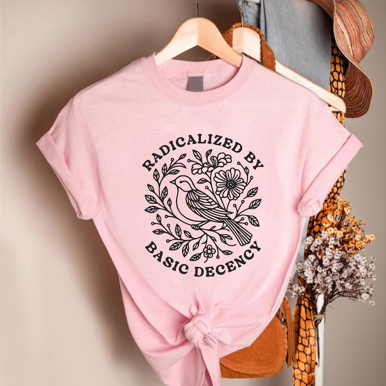 May include: A light pink t-shirt with rolled-up sleeves. The shirt features a black graphic of a bird surrounded by leaves and flowers, with the words "RADICALIZED BY BASIC DECENCY" in a circular design.