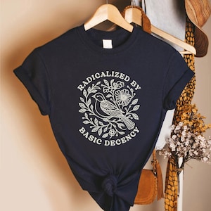 May include: Navy blue t-shirt with rolled-up sleeves. The shirt features a white graphic of a bird, flowers, and leaves, with the text "RADICALIZED BY BASIC DECENCY" in a circular design. The shirt is hanging on a wooden hanger.