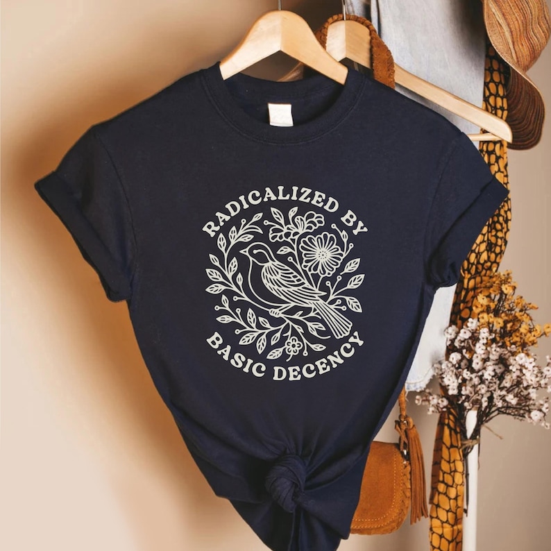 May include: Navy blue t-shirt with rolled-up sleeves. The shirt features a white graphic of a bird, flowers, and leaves, with the text "RADICALIZED BY BASIC DECENCY" in a circular design. The shirt is hanging on a wooden hanger.