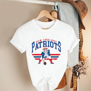May include: White t-shirt featuring the words "New England Patriots" in blue and a graphic of a colonial-era football player. The design includes red and blue stripes, and the shirt is hanging on a wooden hanger.