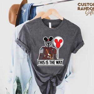 Puede incluir: Camiseta gris oscuro con una ilustraci&oacute;n de dibujos animados de un personaje mandaloriano con un casco tem&aacute;tico de Mickey Mouse y un globo rojo. El texto "THIS IS THE WAY" est&aacute; impreso debajo. La camiseta est&aacute; colgada en una percha de madera.