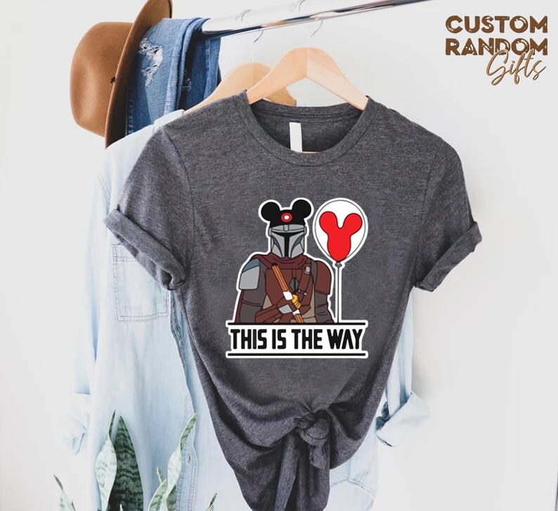Puede incluir: Camiseta gris oscuro con una ilustraci&oacute;n de dibujos animados de un personaje mandaloriano con un casco tem&aacute;tico de Mickey Mouse y un globo rojo. El texto "THIS IS THE WAY" est&aacute; impreso debajo. La camiseta est&aacute; colgada en una percha de madera.
