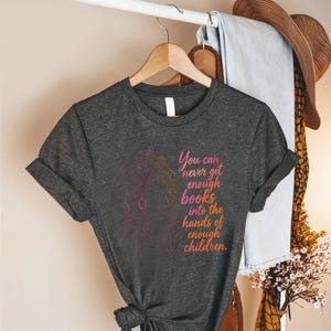 May include: A dark heather gray short-sleeve t-shirt featuring a pink and orange line art illustration of a person with long hair and the text: You can never get enough books into the hands of enough children. The shirt has a crew neckline.