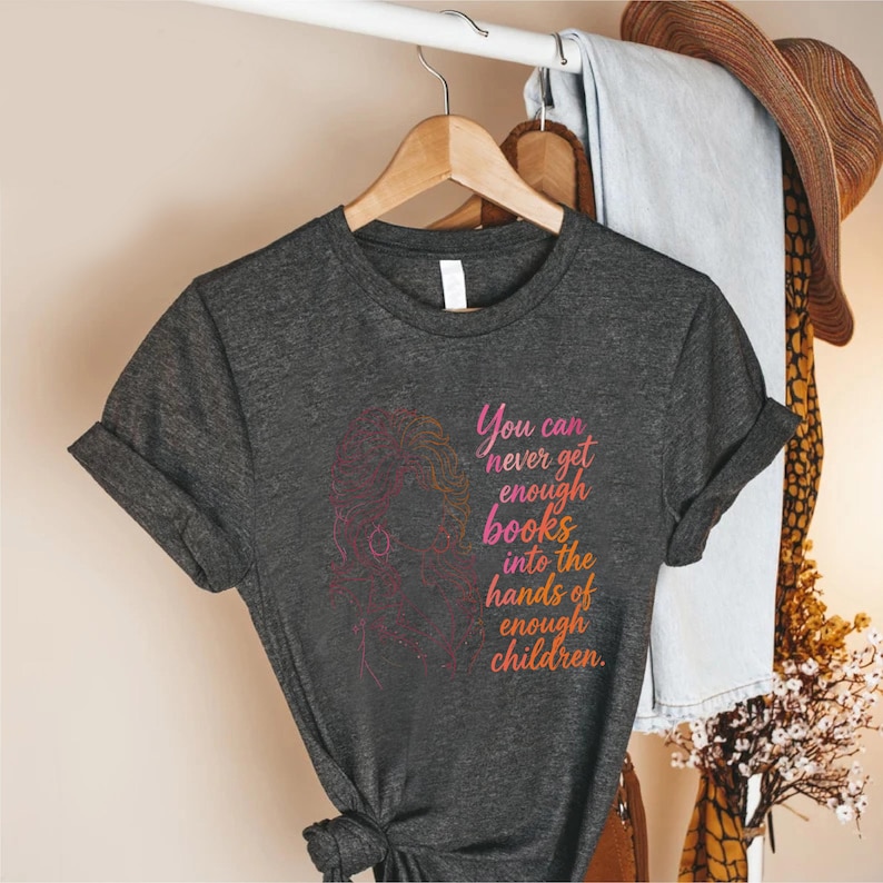 May include: A dark heather gray short-sleeve t-shirt featuring a pink and orange line art illustration of a person with long hair and the text: You can never get enough books into the hands of enough children. The shirt has a crew neckline.