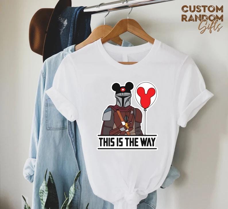 Puede incluir: Camiseta blanca con una ilustraci&oacute;n de dibujos animados de un personaje mandaloriano con una cabeza de Mickey Mouse y un globo rojo. El texto "THIS IS THE WAY" est&aacute; impreso debajo. La camiseta est&aacute; colgada en una percha de madera.