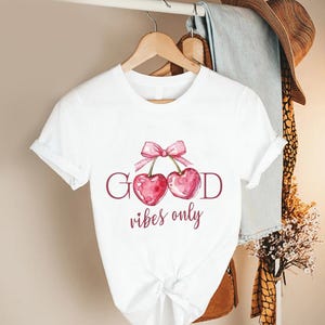 May include: White t-shirt featuring a watercolor design of two cherries with a pink bow and the words "GOOD vibes only". The shirt is hanging on a wooden hanger.