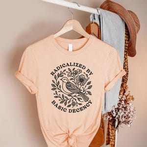 May include: Peach-colored t-shirt with rolled sleeves. The shirt features a black graphic of a bird surrounded by flowers and leaves, with the words "RADICALIZED BY BASIC DECENCY" in a circular design. The shirt is hanging on a wooden hanger.