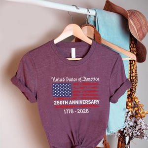 May include: A maroon t-shirt with the text "United States of America" above a stylized American flag design. The flag is red and white with a blue square of white stars. Below the flag is "250TH ANNIVERSARY 1776-2026".
