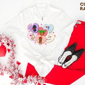 May include: White t-shirt featuring Star Wars characters on Valentine's Day-themed heart-shaped candies. The shirt is paired with red pants and black heels. A Valentine's Day wreath and a necklace are also visible.