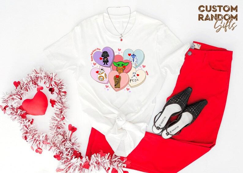 May include: White t-shirt featuring Star Wars characters on Valentine's Day-themed heart-shaped candies. The shirt is paired with red pants and black heels. A Valentine's Day wreath and a necklace are also visible.