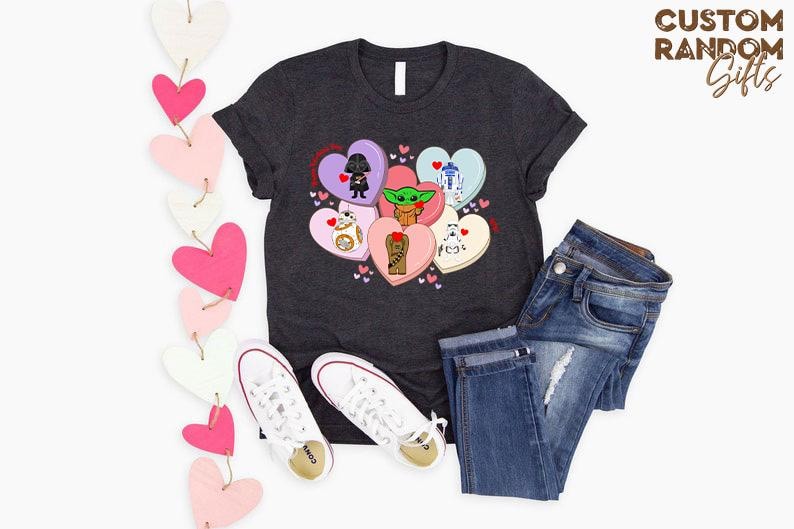 May include: Dark gray t-shirt featuring a heart-shaped graphic with Star Wars characters. The shirt is paired with blue jeans and white sneakers. Decorative pink and white heart garland is on the left. The text "CUSTOM RANDOM Gifts" is in the upper right.