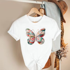May include: White t-shirt featuring a colorful butterfly design. The butterfly is made of patchwork fabric in various colors, including red, blue, green, and pink. The shirt has short sleeves and a round neckline.