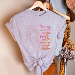 May include: A heather gray short-sleeved t-shirt featuring a pink and orange gradient line art illustration of a woman with voluminous hair and the text, You can never get enough books into the hands of enough children, printed on the front.