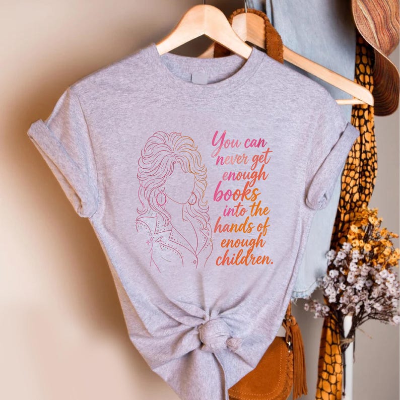 May include: A heather gray short-sleeved t-shirt featuring a pink and orange gradient line art illustration of a woman with voluminous hair and the text, You can never get enough books into the hands of enough children, printed on the front.