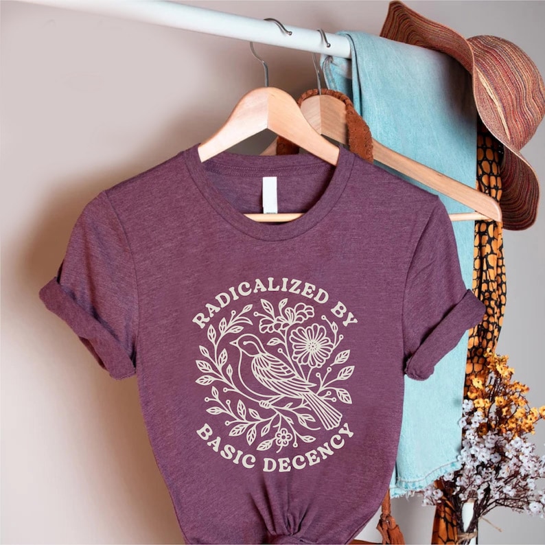 May include: A maroon t-shirt with rolled-up sleeves. The shirt features a white graphic of a bird, flowers, and leaves, with the text "RADICALIZED BY BASIC DECENCY" in a circular design. The shirt is hanging on a wooden hanger.