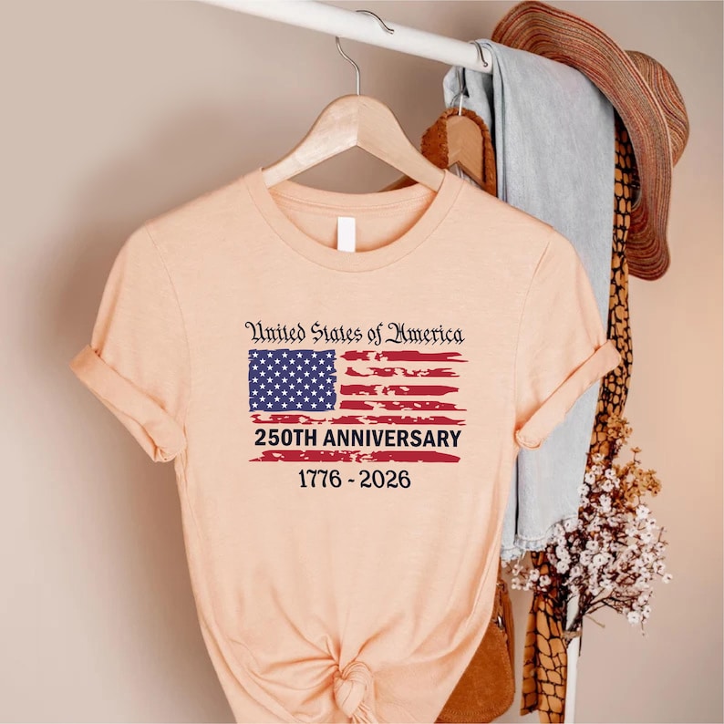May include: A peach-colored t-shirt with the text "United States of America" above a stylized American flag. The flag design includes a blue square with white stars and red stripes. Below the flag, it reads "250TH ANNIVERSARY 1776-2026".