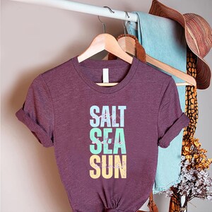 May include: A maroon t-shirt with rolled-up sleeves. The shirt features the words "SALT SEA SUN" in a vertical stack, with each word in a different color: light blue, seafoam green, and yellow. The shirt is hanging on a wooden hanger.