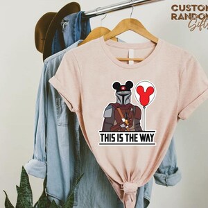 Puede incluir: Camiseta rosa claro con una ilustraci&oacute;n de dibujos animados de un personaje mandaloriano con un casco tem&aacute;tico de Mickey Mouse y un globo rojo. El texto "THIS IS THE WAY" est&aacute; impreso debajo.