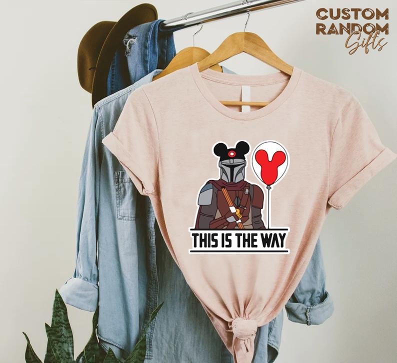 Puede incluir: Camiseta rosa claro con una ilustraci&oacute;n de dibujos animados de un personaje mandaloriano con un casco tem&aacute;tico de Mickey Mouse y un globo rojo. El texto "THIS IS THE WAY" est&aacute; impreso debajo.