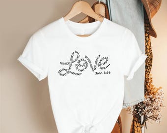 May include: White t-shirt with the text "LOVE" in a circular design, featuring the words "For God so loved the world..." and "John 3:16". The shirt is hanging on a wooden hanger.