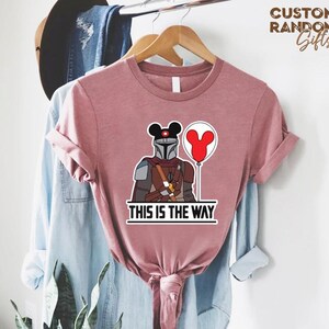Puede incluir: Camiseta rosa polvoriento con un gr&aacute;fico de un personaje mandaloriano con un casco de orejas de Mickey Mouse y un globo rojo. El texto "THIS IS THE WAY" est&aacute; impreso debajo. La camiseta est&aacute; colgada en una percha de madera.