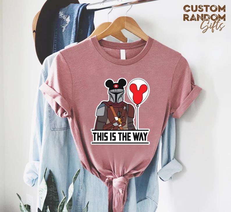 Puede incluir: Camiseta rosa polvoriento con un gr&aacute;fico de un personaje mandaloriano con un casco de orejas de Mickey Mouse y un globo rojo. El texto "THIS IS THE WAY" est&aacute; impreso debajo. La camiseta est&aacute; colgada en una percha de madera.