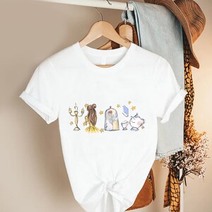 May include: White t-shirt featuring a whimsical illustration of a candelabra, a woman's hairstyle, a rose in a glass dome, a teapot, and other elements. The design is in soft pastel colors with gold accents.