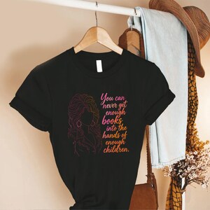 May include: A black short-sleeved t-shirt featuring a pink and orange gradient line art illustration of a person with voluminous hair. The text reads: You can never get enough books into the hands of enough children. The shirt is displayed on a hanger.