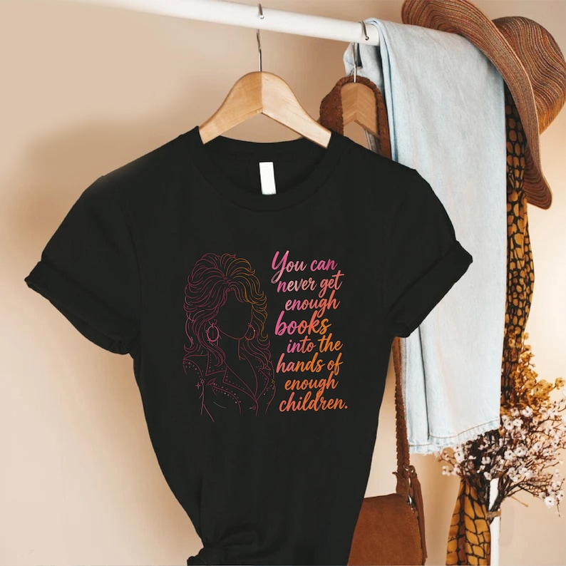 May include: A black short-sleeved t-shirt featuring a pink and orange gradient line art illustration of a person with voluminous hair. The text reads: You can never get enough books into the hands of enough children. The shirt is displayed on a hanger.