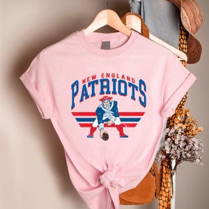 May include: A light pink t-shirt featuring the words "NEW ENGLAND PATRIOTS" in blue lettering. Below the text is a graphic of a colonial-era figure in a blue coat and red pants holding a football. Red and blue stripes are behind the figure.
