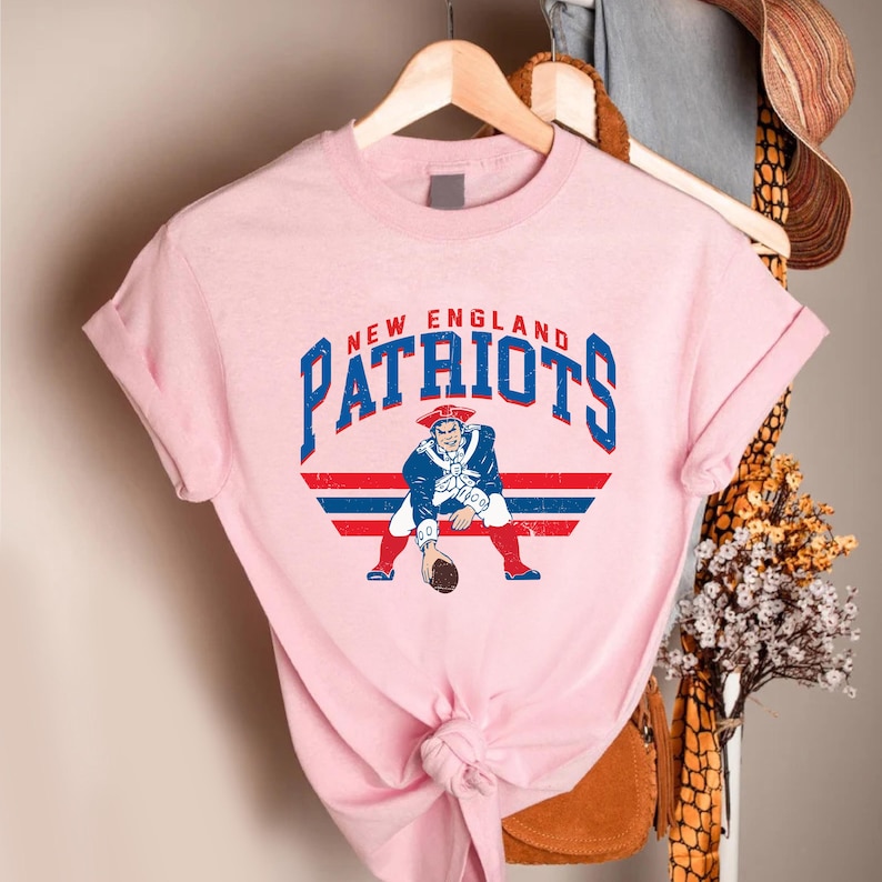 May include: A light pink t-shirt featuring the words "NEW ENGLAND PATRIOTS" in blue lettering. Below the text is a graphic of a colonial-era figure in a blue coat and red pants holding a football. Red and blue stripes are behind the figure.