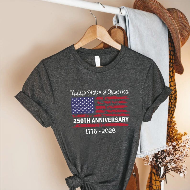 May include: Dark gray t-shirt featuring a distressed American flag design with the text "United States of America" and "250th Anniversary 1776-2026". The shirt is hanging on a wooden hanger.