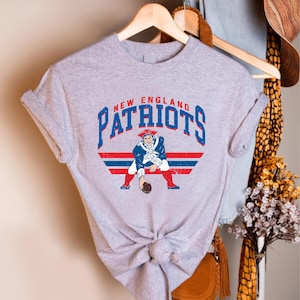 May include: A heather gray t-shirt featuring the words "NEW ENGLAND PATRIOTS" in blue and red arched text. Below the text is a graphic of a colonial-era figure in a blue coat, red pants, and a tricorn hat, holding a football.