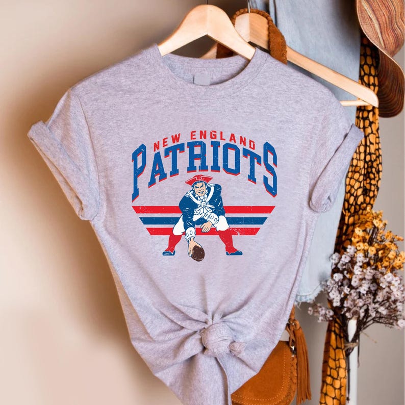 May include: A heather gray t-shirt featuring the words "NEW ENGLAND PATRIOTS" in blue and red arched text. Below the text is a graphic of a colonial-era figure in a blue coat, red pants, and a tricorn hat, holding a football.